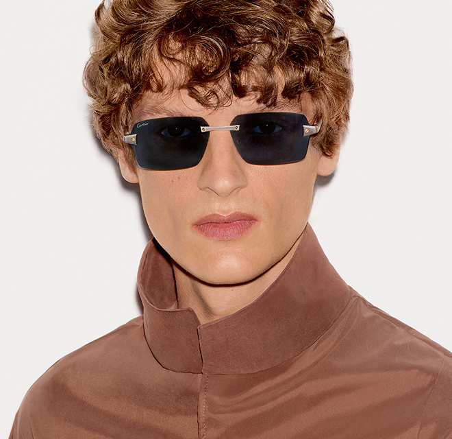 A man in a brown jacket and sunglasses stands confidently, showcasing a stylish outdoor look.