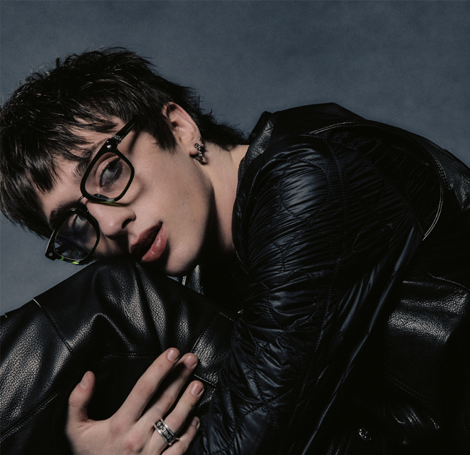 A man with glasses wearing a black leather jacket, standing confidently with a neutral expression.